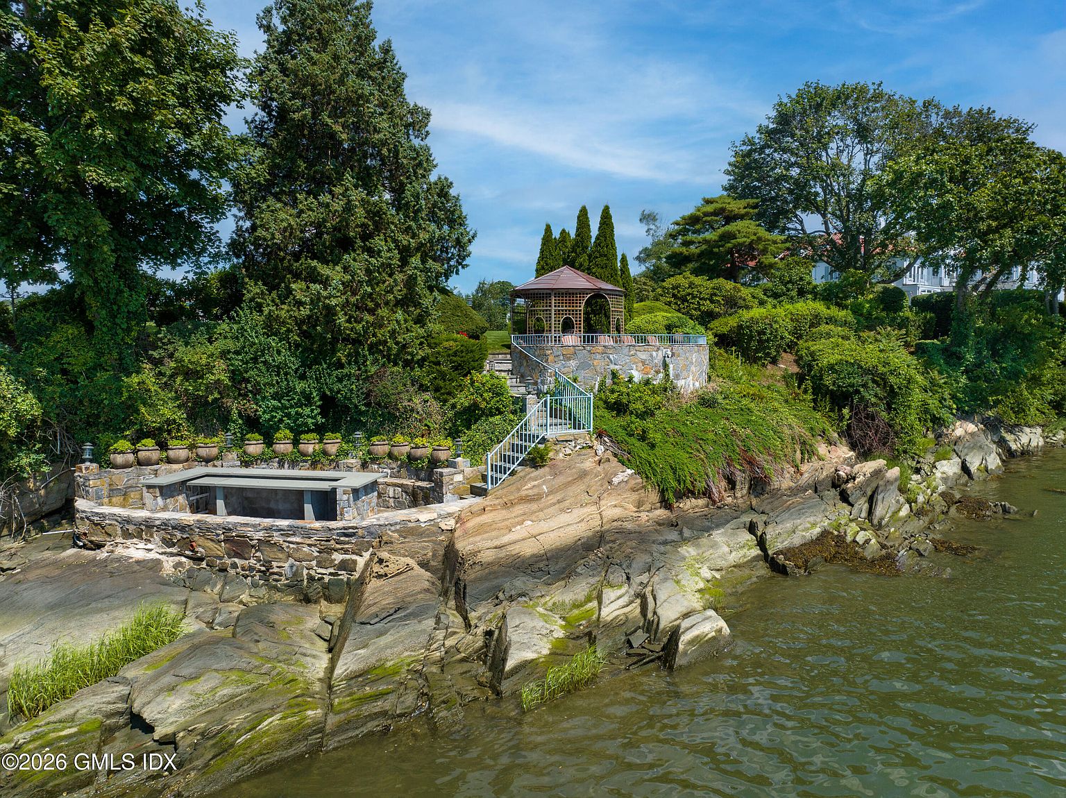This elevated aerial perspective captures a scenic waterfront garden featuring a stone-walled gazebo perched on a rocky outcrop. A stone-built outdoor seating area with a long bench sits lower on the rocks, bordered by a row of decorative planters. The landscape is lush with mature trees and greenery, creating a private, tranquil retreat overlooking the water.