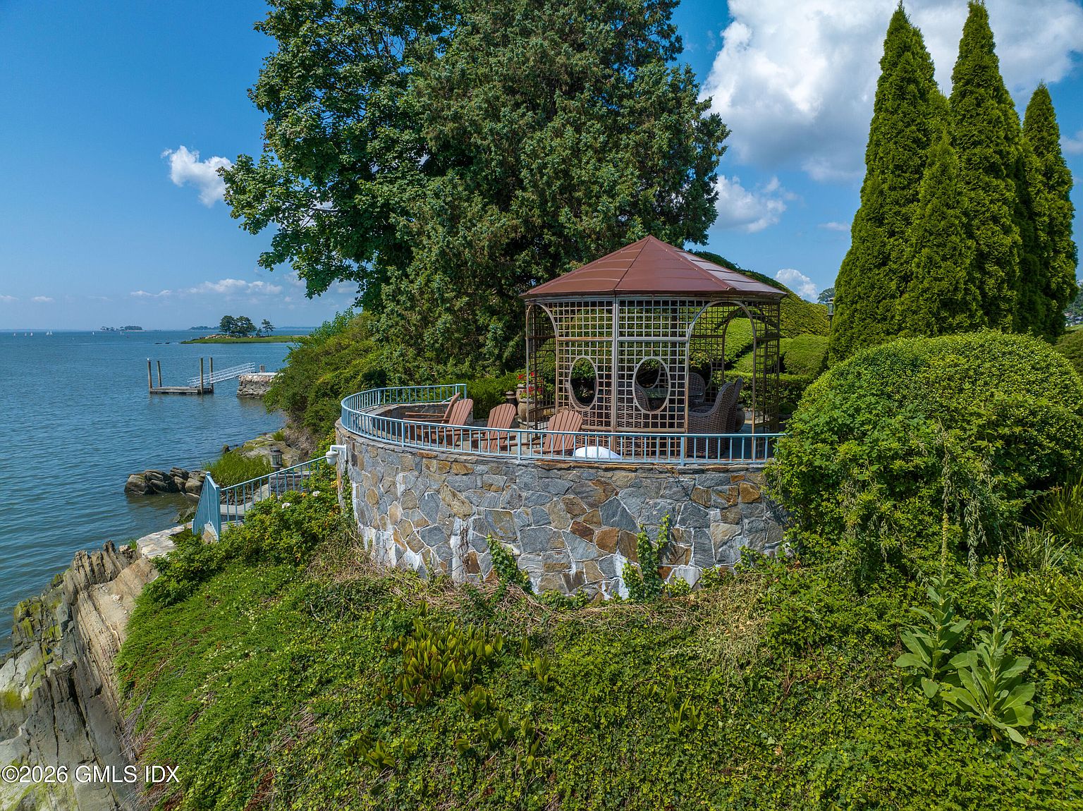 This elevated perspective captures a scenic waterfront garden featuring a stone-walled terrace topped with a metal-roofed gazebo. The space is furnished with outdoor seating, offering expansive views of the adjacent bay and a private dock. Lush greenery, including tall evergreen trees and dense shrubs, frames the area, creating a serene and private coastal retreat.