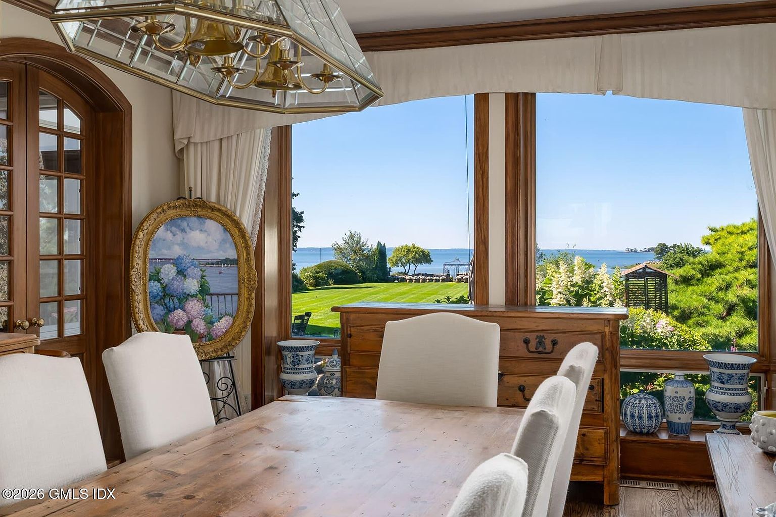 This elegant dining room features a large wooden table surrounded by upholstered chairs, positioned to take full advantage of expansive windows overlooking a lush green lawn and the water beyond. A prominent oval painting of hydrangeas rests against the window frame, while a large, multi-tiered brass chandelier hangs above the table. The room is accented with blue and white decorative pottery, creating a sophisticated and serene atmosphere with a strong connection to the outdoor landscape.