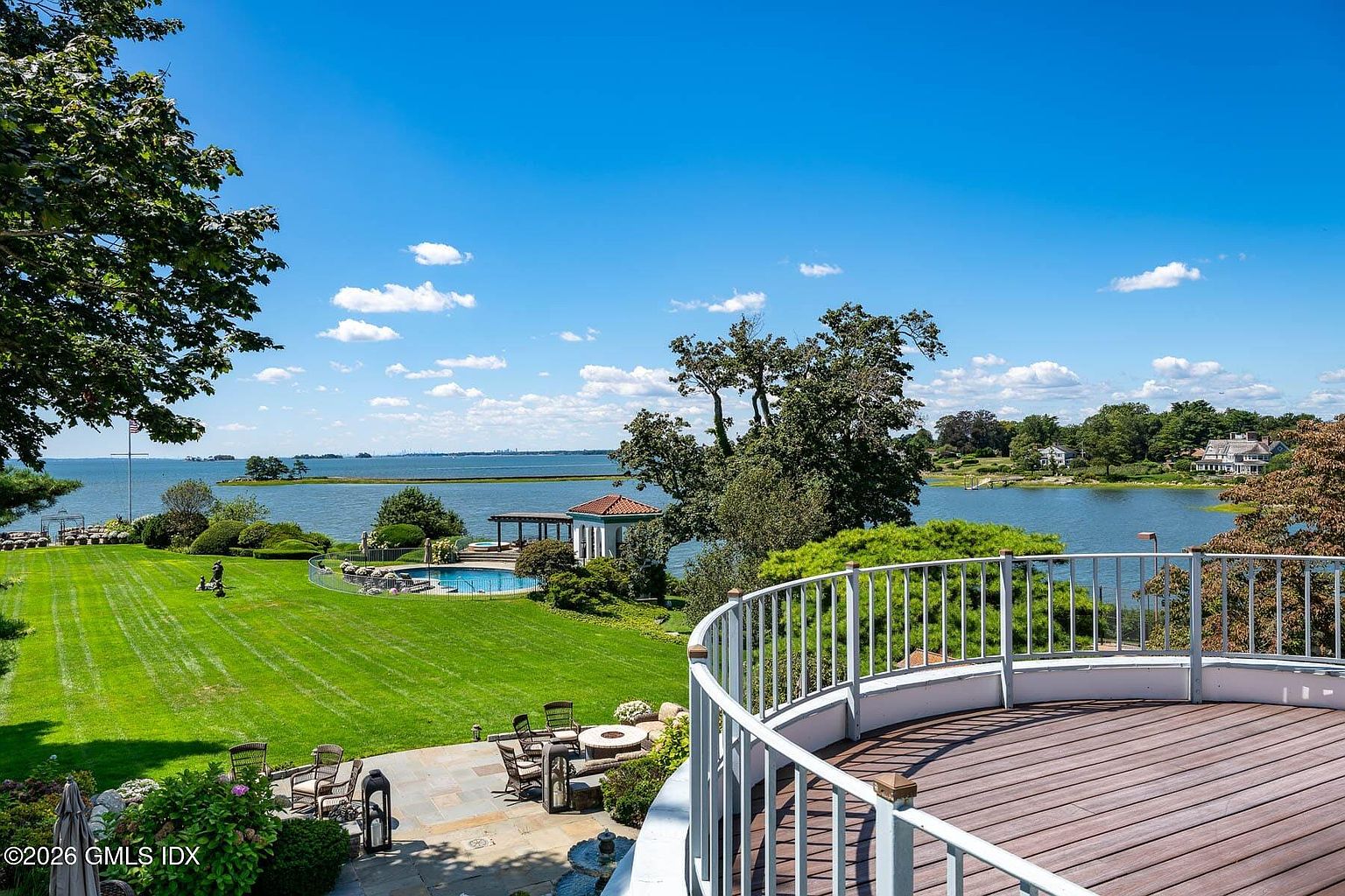 This elevated perspective captures a sweeping view from a curved wooden deck overlooking a lush, expansive lawn that leads down to a serene waterfront. The scene features a private swimming pool with a nearby gazebo, set against a backdrop of mature trees and a clear blue sky. The vantage point emphasizes the property's prime location and seamless transition between outdoor living spaces and the natural landscape.