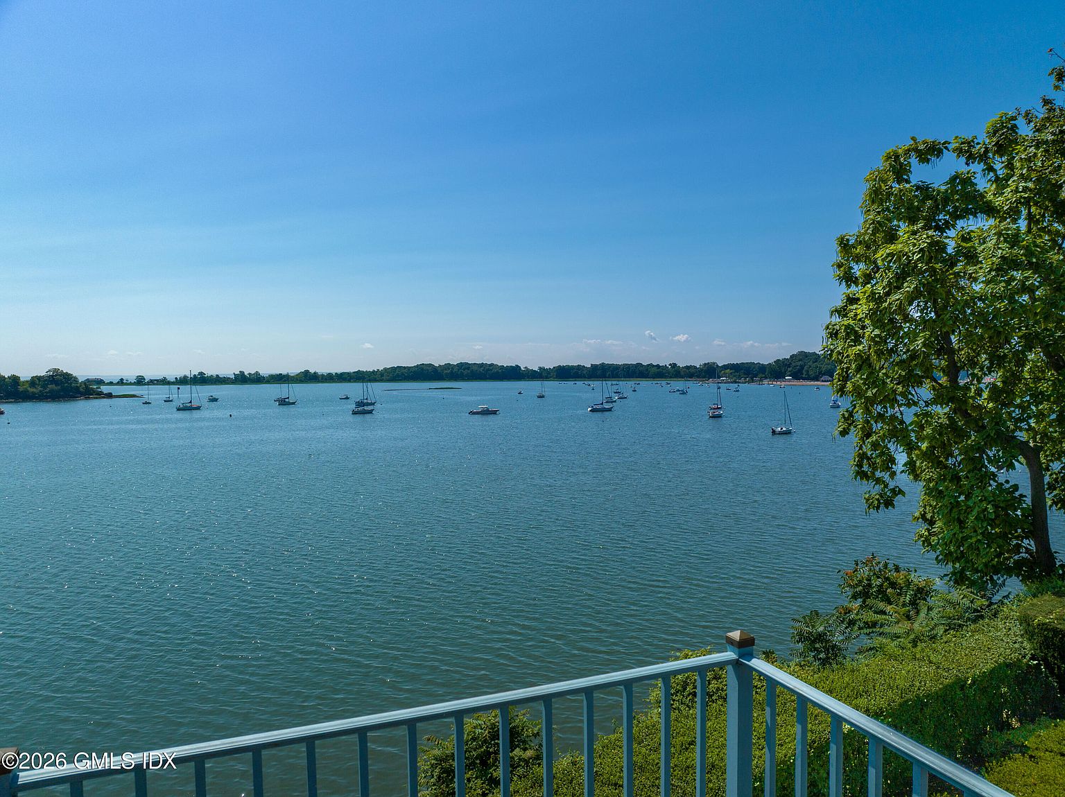 This image captures a scenic, elevated view from a balcony overlooking a calm, expansive bay filled with numerous sailboats. The foreground features a blue metal railing, while a large, leafy tree frames the right side of the composition. The perspective provides a serene, wide-angle look at the waterfront, emphasizing the property's prime location and tranquil atmosphere.