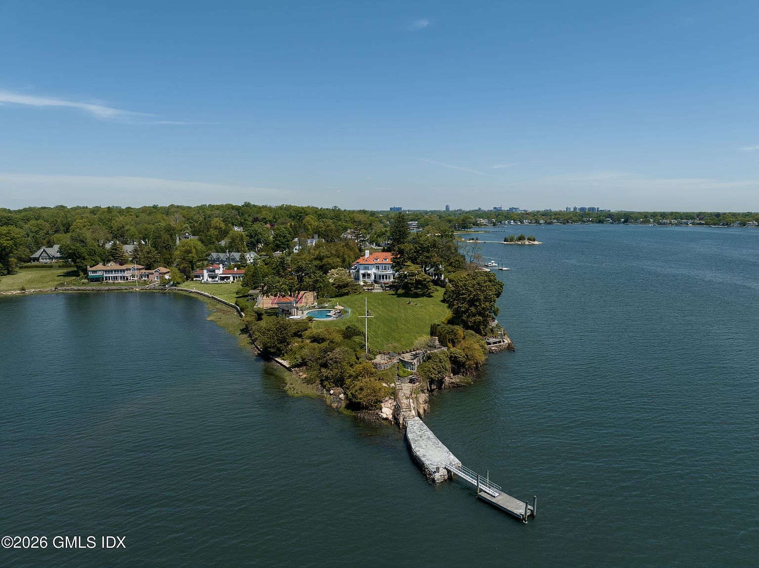 This high-angle aerial perspective showcases a sprawling waterfront estate situated on a private peninsula surrounded by calm water. The property features a grand residence with a red-tiled roof, a circular swimming pool, and a long stone pier extending into the bay. Lush green lawns and mature trees frame the home, creating a serene and exclusive coastal atmosphere.