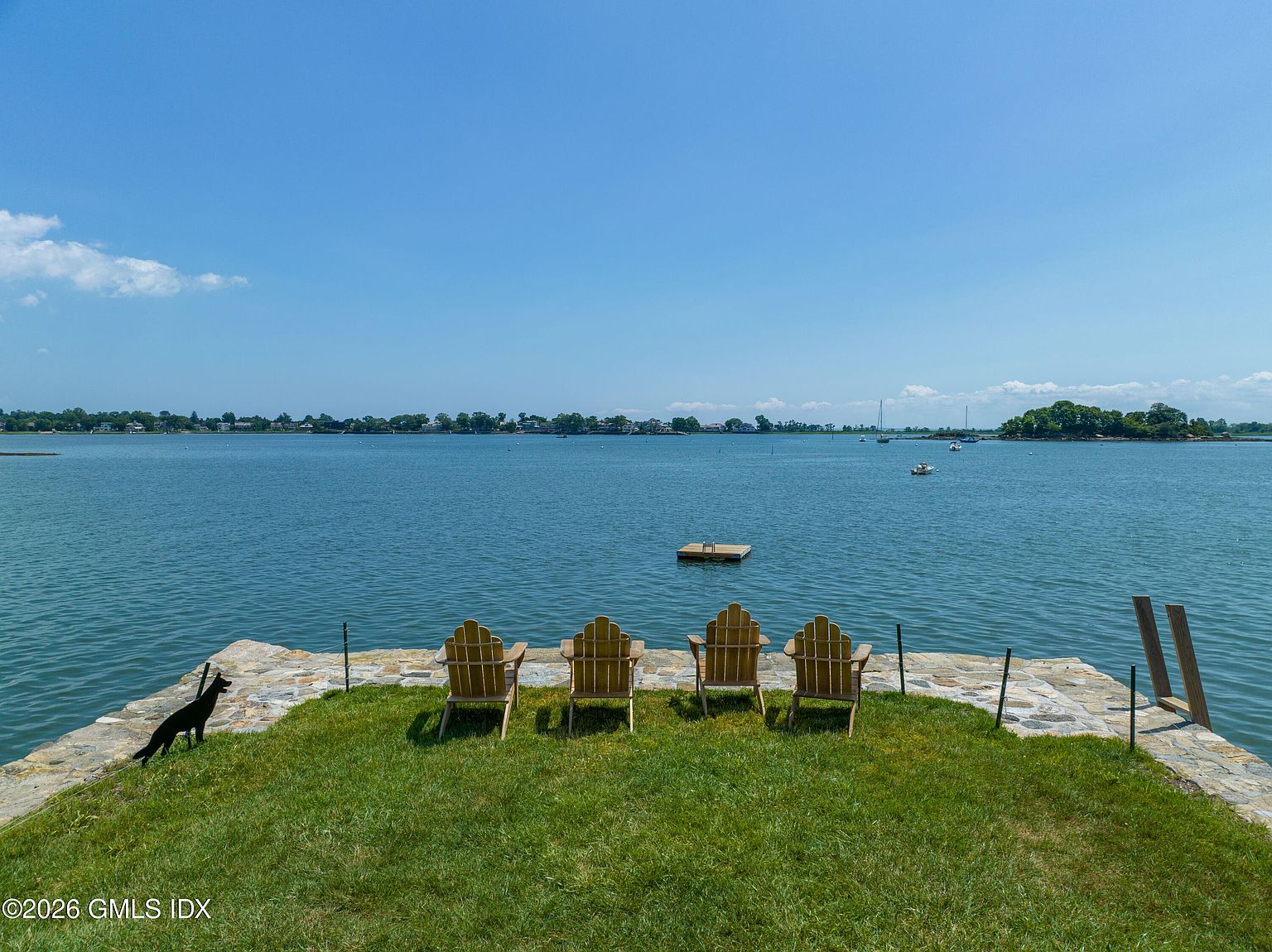 This scenic waterfront property features a stone-paved terrace overlooking a calm bay, furnished with four wooden Adirondack chairs. A lush green lawn leads directly to the water's edge, where a small floating dock sits in the distance. The wide-angle perspective captures the expansive blue sky and the tranquil coastal horizon, creating a serene and inviting outdoor living space.
