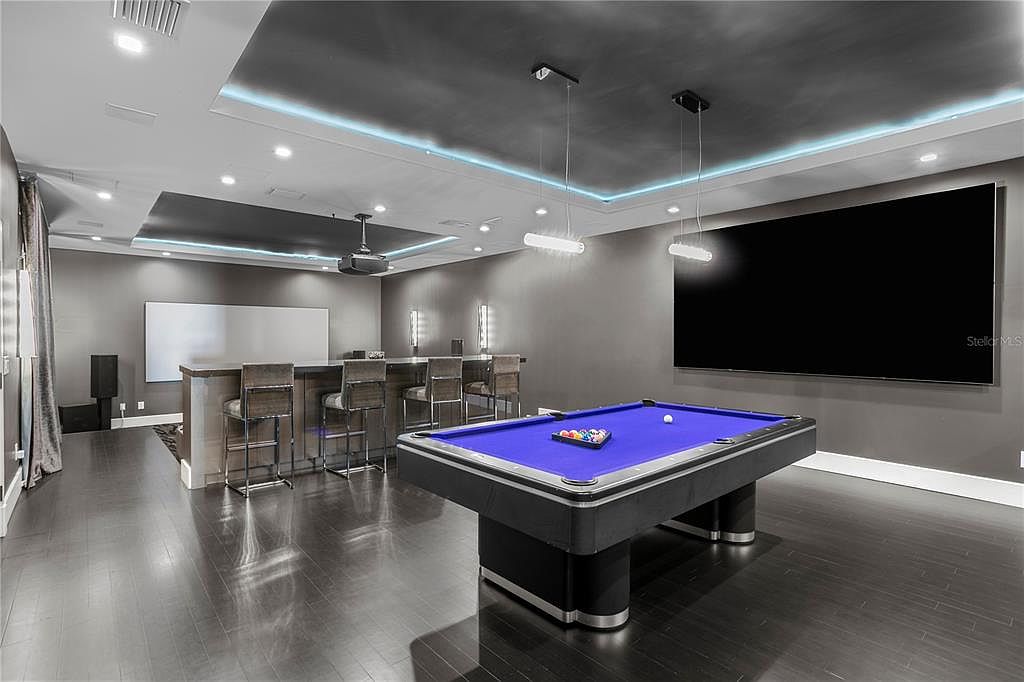 This is a finished basement featuring a pool table, a large screen, and a bar area with seating. The room has dark hardwood floors and gray walls, creating a modern and entertainment-focused space. Recessed lighting and pendant lights add to the ambiance.