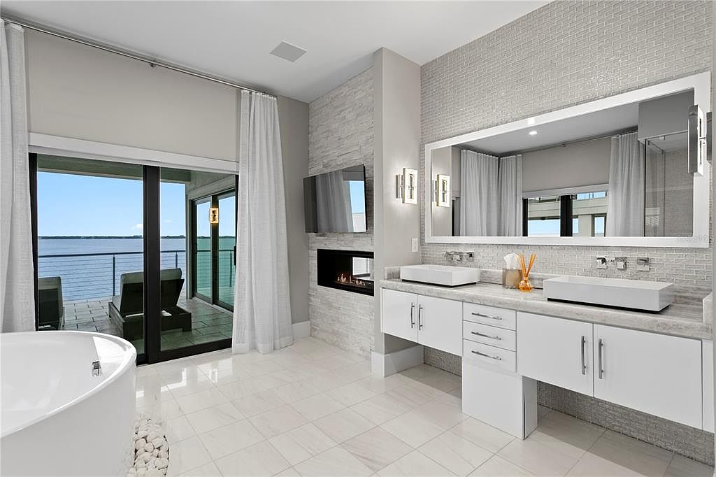 This is a luxurious primary bathroom featuring a double vanity with white cabinetry and marble countertops, complemented by a large mirror and modern lighting fixtures. A freestanding bathtub sits near a sliding glass door that opens to a balcony with water views, and a fireplace with a TV above adds a touch of elegance. The overall impression is one of spa-like tranquility and modern sophistication.