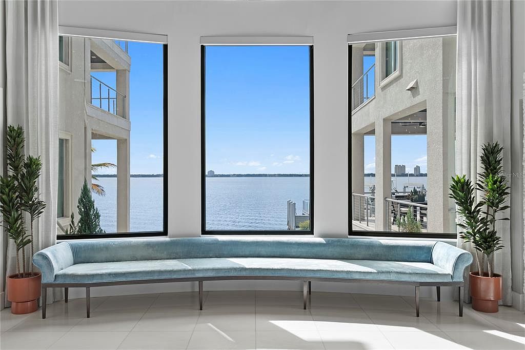 This interior shot showcases a modern living room with a long, curved blue velvet sofa positioned in front of three large windows offering a scenic water view. The room is bright and airy, featuring neutral-toned walls, tile flooring, and potted plants on either side of the sofa, creating a serene and stylish atmosphere.