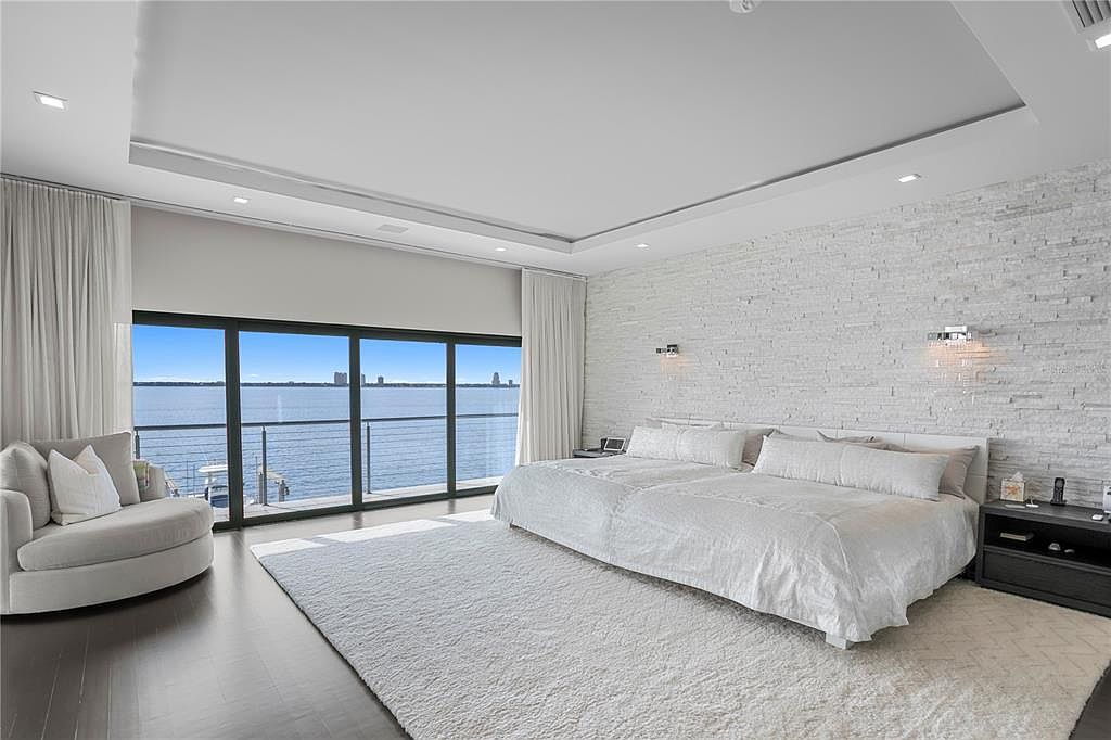 This is a bright and spacious primary bedroom with a modern aesthetic. The room features a large bed with white linens, a textured white brick wall, and floor-to-ceiling windows offering a waterfront view. A comfortable lounge chair and a plush white rug add to the luxurious feel of the space.