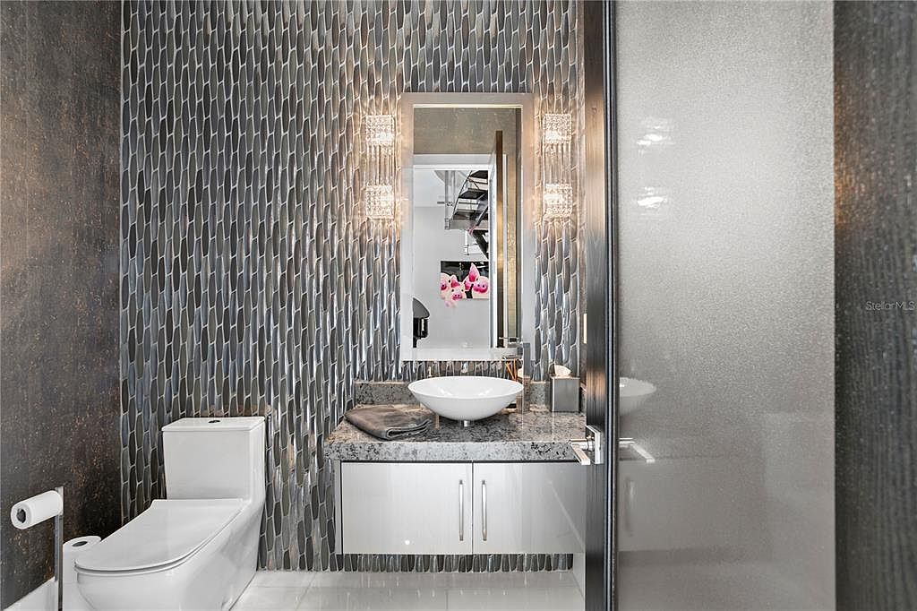 This is a modern guest bathroom featuring a unique textured wall, a vessel sink on a granite countertop, and a sleek white vanity. The room is illuminated by decorative sconces flanking a mirror, and a toilet is visible to the left. The overall design is stylish and contemporary, creating a luxurious feel for guests.