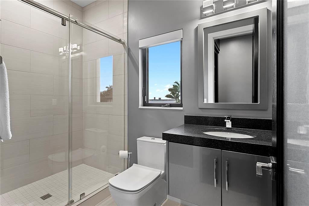 This is a modern bathroom featuring a glass-enclosed shower with light beige tiling, a white toilet, and a gray vanity with a black countertop. A window provides natural light, and the overall color scheme is neutral and contemporary. The perspective is from the doorway, showcasing the entire bathroom layout.