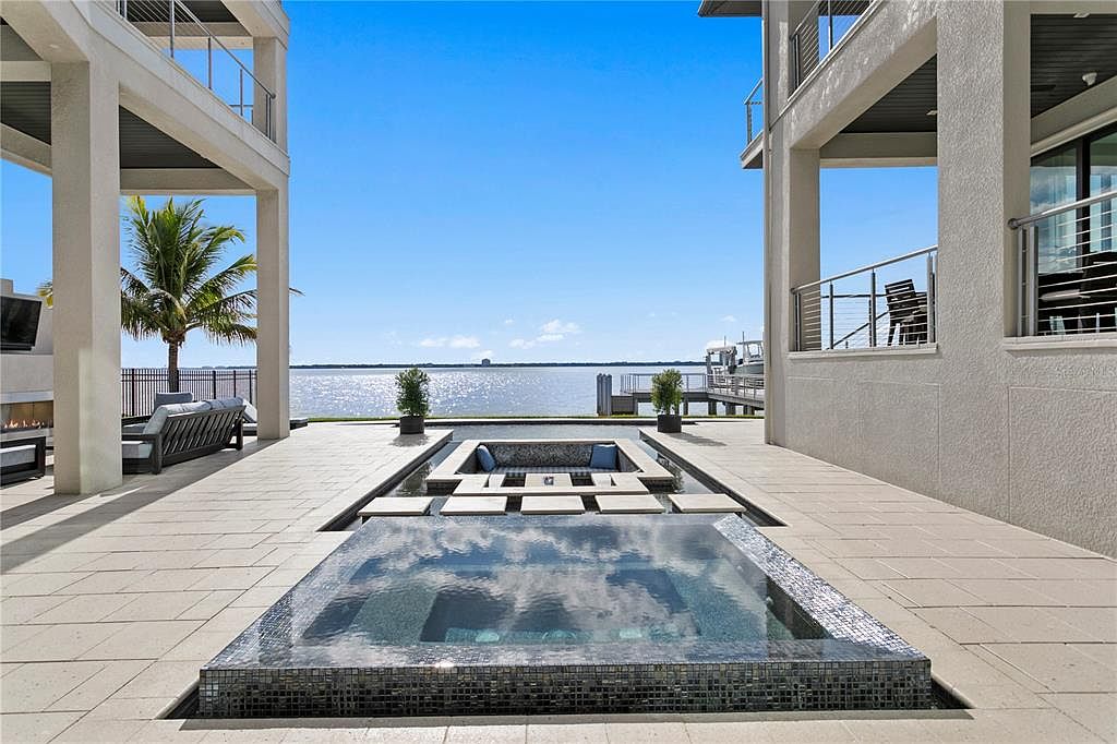 This exterior shot showcases a luxurious pool and spa area with a stunning waterfront view. The modern design features a tiled pool and spa, surrounded by a spacious patio with elegant outdoor furniture and landscaping. The clear blue sky and calm water create a serene and inviting atmosphere, perfect for relaxation and entertainment.