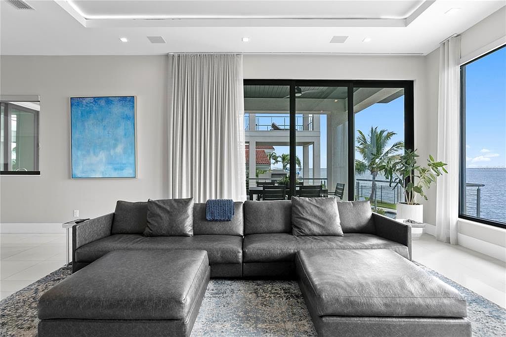 This is an interior shot of a modern living room featuring a large, gray sectional sofa with matching ottomans, set upon a patterned area rug. A blue abstract painting hangs on the wall, and floor-to-ceiling windows and sliding glass doors provide ample natural light and a view of the outdoor patio and waterfront. The room exudes a contemporary and luxurious feel.