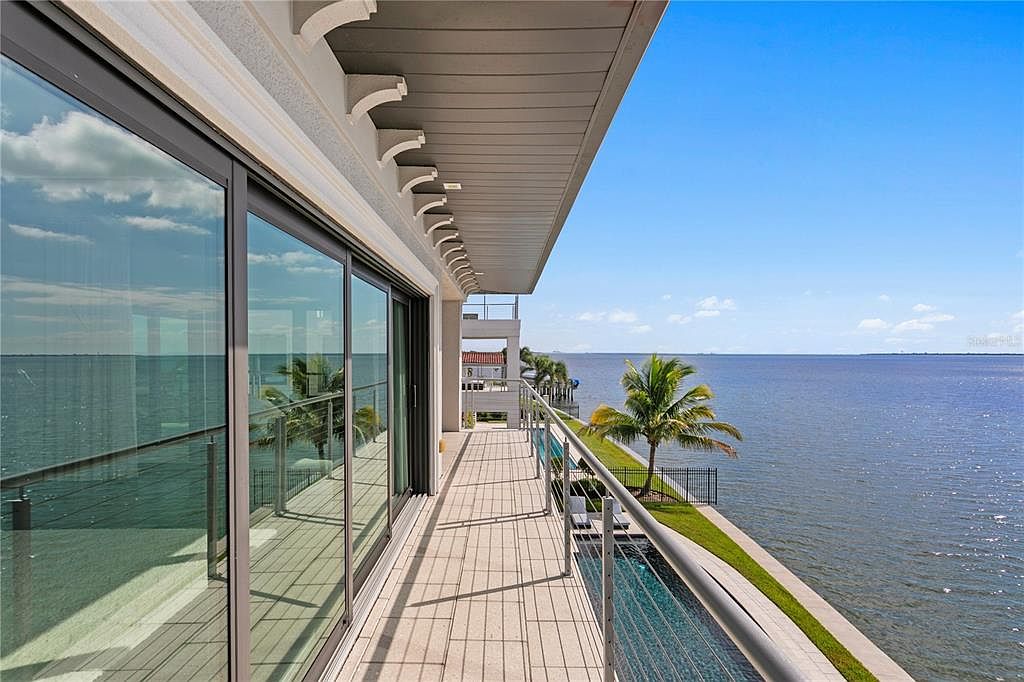 This image showcases a luxurious waterfront balcony with a sleek, modern design. The balcony features a tiled floor, a glass railing, and sliding glass doors that lead into the interior. The view overlooks a serene body of water, with a palm tree and a glimpse of a pool adding to the upscale ambiance.