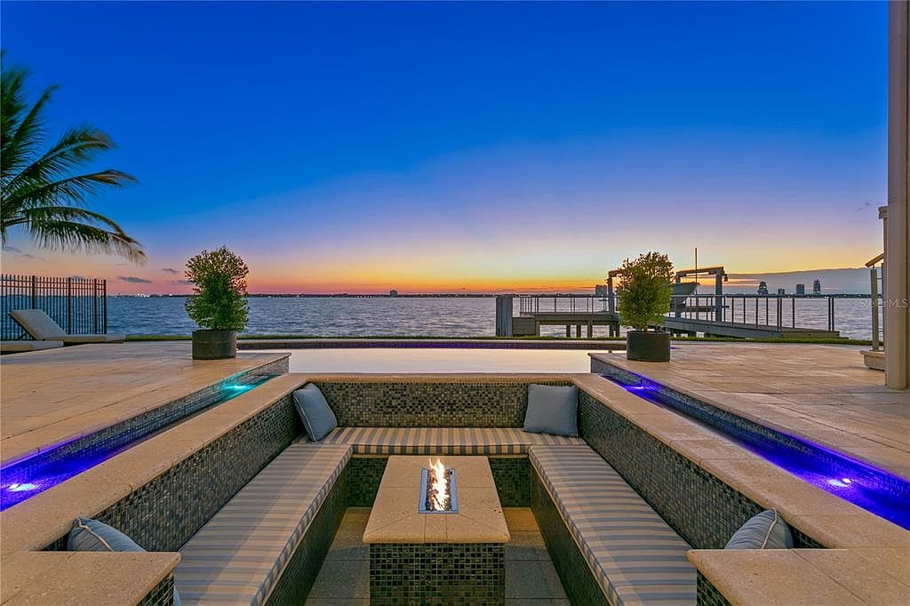 This image showcases a luxurious outdoor patio area at dusk, featuring a built-in seating arrangement with plush cushions and a central fire pit. The patio overlooks a serene body of water, with a dock and distant city skyline visible in the background. The scene is enhanced by subtle blue lighting along the edges of the patio, creating a sophisticated and inviting ambiance.