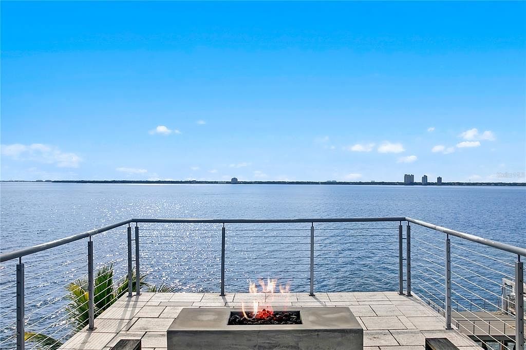 This image showcases a modern patio or deck overlooking a serene body of water, possibly a lake or ocean. The space features a contemporary fire pit as a focal point, surrounded by sleek railings and a tiled surface. The view extends to the horizon, offering a sense of tranquility and luxury, perfect for outdoor relaxation and entertainment.