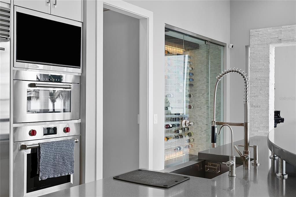 This is a modern kitchen featuring stainless steel appliances, including a built-in oven and microwave. The kitchen island has a sleek countertop and a modern faucet. A glass-enclosed wine cellar is visible in the background, adding a touch of luxury to the space.