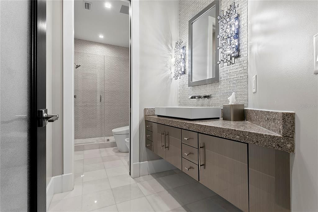 This is a modern bathroom featuring a floating vanity with a light brown finish and a speckled countertop. A rectangular vessel sink sits atop the counter, complemented by a sleek faucet and a decorative mirror with sconces. The bathroom also includes a walk-in shower with glass doors and tiled walls, creating a clean and contemporary aesthetic.