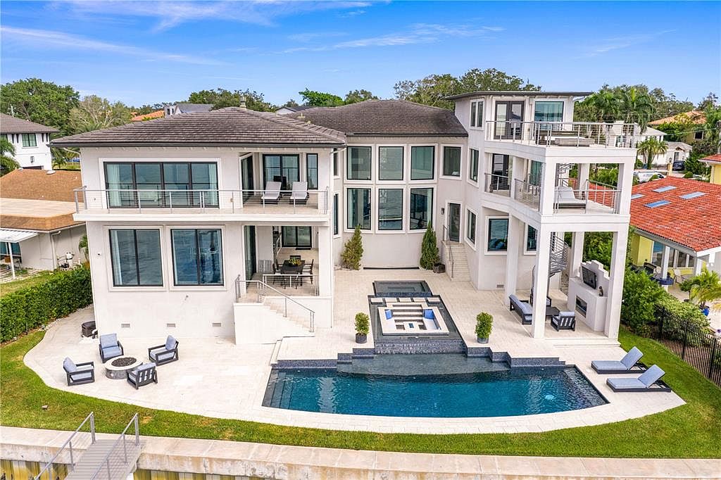 This is a rear exterior view of a luxurious multi-story home featuring a modern design with white stucco and dark-framed windows. The property includes multiple balconies, a swimming pool, a spa, and an outdoor seating area with a fire pit. The landscaping is well-maintained, and the overall impression is one of high-end living and sophisticated outdoor entertainment.