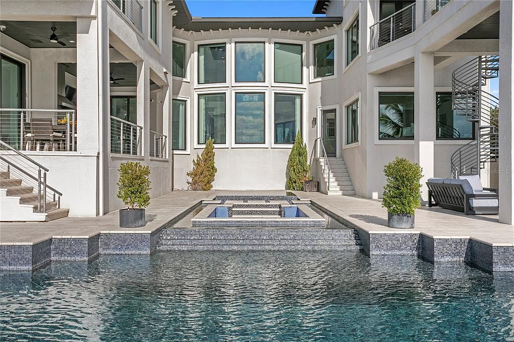 This image showcases a luxurious pool and spa area in the rear of a modern home. The pool features dark blue water and a raised spa with intricate tile work. The architecture includes multiple levels, balconies, and a spiral staircase, creating a sophisticated outdoor living space.