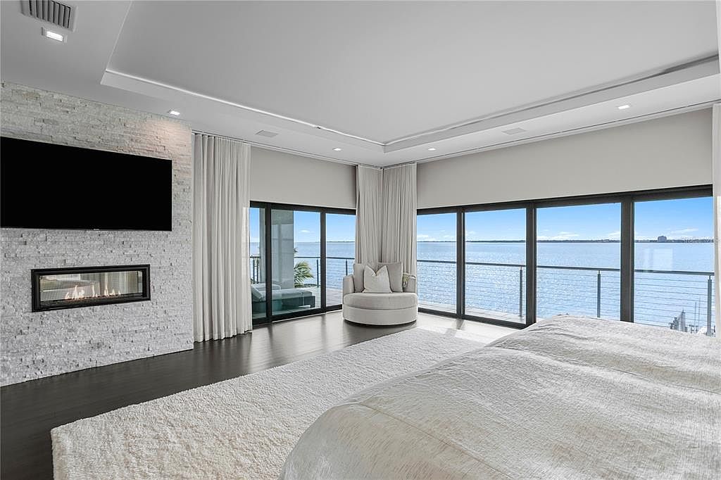 This is a luxurious primary bedroom featuring a modern fireplace with a large television above it, floor-to-ceiling windows with water views, and a comfortable seating area. The room is decorated in neutral tones with a large white rug and dark wood floors, creating a serene and upscale atmosphere. The perspective is from the foot of the bed, looking towards the windows and seating area.