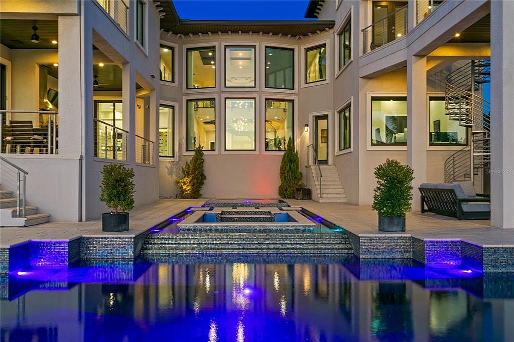 This is a stunning rear exterior view showcasing a luxurious pool and spa area of a modern home. The pool features vibrant blue lighting, reflecting the surrounding architecture and creating a dramatic effect. The design incorporates tiered levels with mosaic tile accents, complemented by lush greenery and architectural lighting, enhancing the property's appeal and creating an inviting outdoor space.