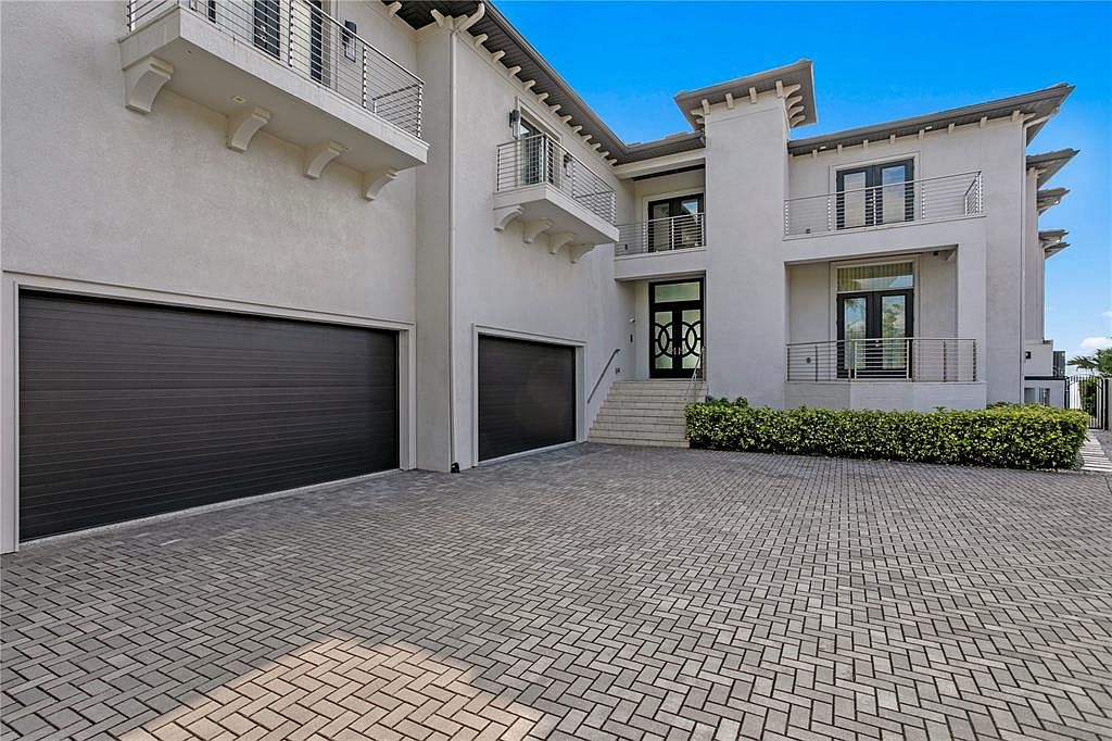 This is a front exterior view of a modern two-story home. The house features a light-colored facade, dark garage doors, and balconies with metal railings. The driveway is paved with interlocking bricks, and there is manicured greenery near the entrance, creating a welcoming and sophisticated curb appeal.