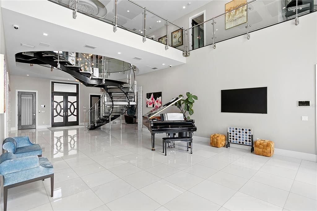 This grand living room features a soaring two-story ceiling, a striking spiral staircase, and glossy white tile flooring that reflects ample natural light. A black grand piano adds a touch of elegance, while modern furnishings and a large flat-screen TV provide comfort and entertainment. The space exudes luxury and sophistication, perfect for both relaxation and entertaining.