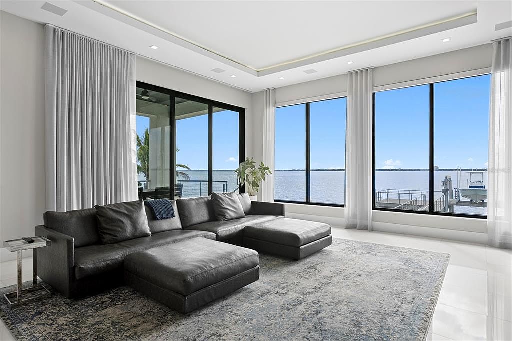 This is a spacious living room featuring a large, dark gray sectional sofa and a patterned area rug. Floor-to-ceiling windows offer expansive water views, enhancing the room's bright and airy atmosphere. The room is decorated in a modern style with neutral tones, creating a luxurious and comfortable living space.