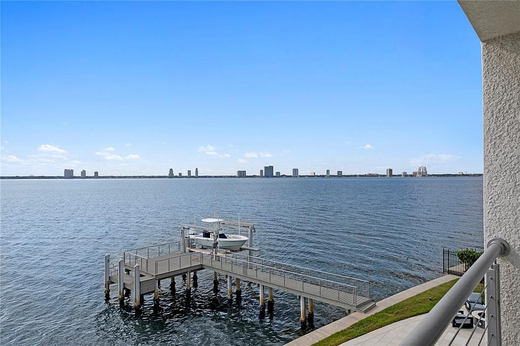 This image showcases a waterfront property from a balcony perspective, featuring a private dock with a boat lift and a boat. The view extends across the water to a distant cityscape under a clear blue sky. The scene conveys a sense of luxury and waterfront living.