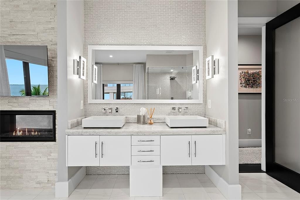 This is a well-lit primary bathroom featuring a double vanity with white cabinetry and marble countertops. A large mirror is centered above the sinks, flanked by modern sconces. The bathroom has a clean, contemporary design with a neutral color palette and a glimpse into an adjacent room through a doorway.