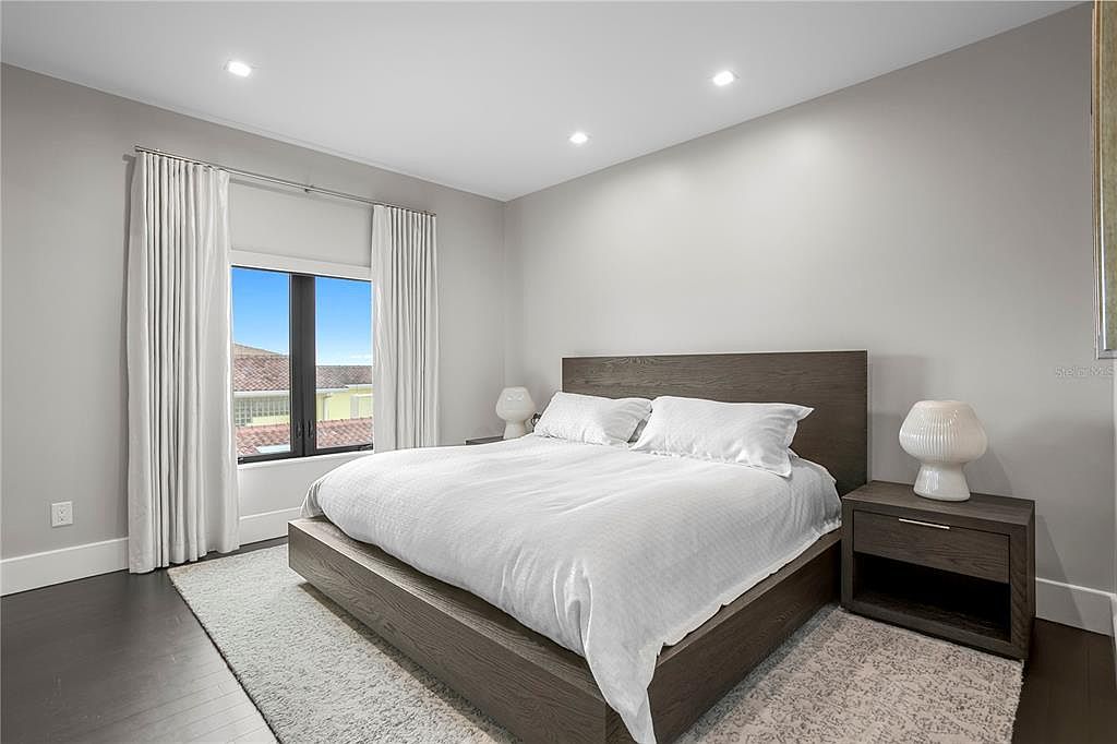 This is a well-lit primary bedroom featuring a large bed with white linens and a dark wood frame, flanked by matching nightstands with modern lamps. The room has a neutral color palette with light gray walls, dark wood flooring, and white curtains framing a window with a view of the outside. The perspective is from the foot of the bed, showcasing the entire sleeping area.
