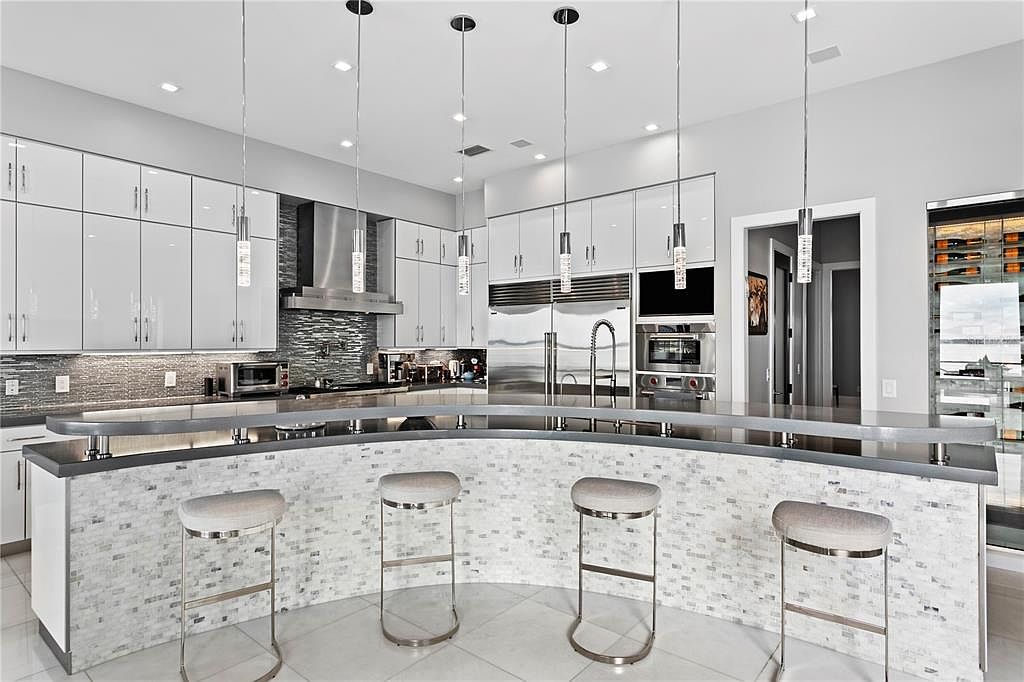 This is a modern kitchen with white, glossy cabinets and stainless steel appliances. A curved island with a mosaic tile base and a dark countertop is the focal point, complemented by four bar stools. Pendant lights hang above the island, adding to the contemporary aesthetic.