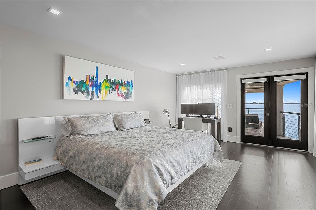 This is a primary bedroom featuring a modern design with a large bed, a cityscape painting above the headboard, and a desk area. The room has dark hardwood floors, a gray area rug, and double doors leading to a balcony with a water view. The overall impression is luxurious and serene.