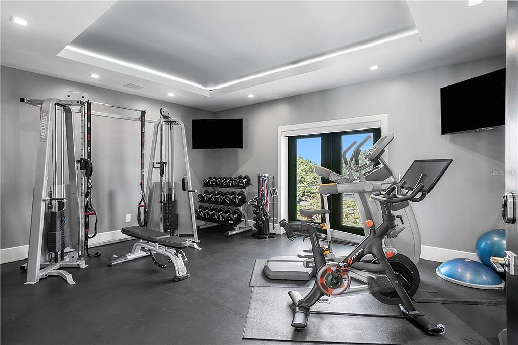 This is a well-equipped home gym featuring weight machines, free weights, and cardio equipment like a Peloton bike and elliptical. The room has a sleek, modern design with gray walls, black flooring, and recessed lighting. A large window provides natural light and a view to the outside, enhancing the workout environment.