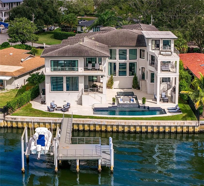 This is an exterior rear view of a luxurious waterfront home featuring a modern design with multiple balconies and large windows. The property includes a swimming pool, a fire pit area, and a private dock with a boat lift, offering direct access to the water. The landscaping is well-maintained, enhancing the overall appeal of this high-end residence.