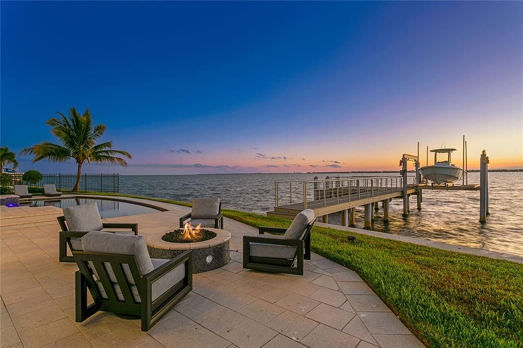 This exterior shot showcases a luxurious patio area with comfortable seating around a modern fire pit, offering a serene waterfront view. A private dock extends into the water, complete with a boat lift, enhancing the property's appeal for boating enthusiasts. The scene is set against a beautiful twilight sky, creating an inviting and upscale ambiance.