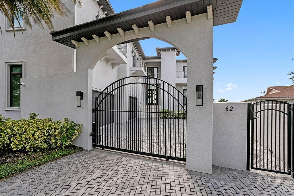This image showcases the grand entryway of a modern home, featuring a black wrought iron gate set within a stucco archway. The address '82' is prominently displayed on the right pillar. The herringbone brick driveway leads to the gate, and lush greenery adds a touch of nature to the scene, creating an inviting and secure entrance.