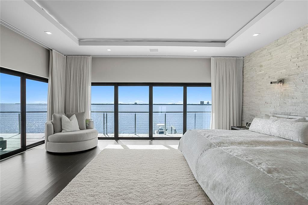This is a luxurious primary bedroom featuring a king-size bed with white linens, a textured accent wall, and floor-to-ceiling windows offering stunning water views. A round chair sits near the windows, and a plush rug covers the dark wood floor. The room exudes a serene and upscale ambiance.