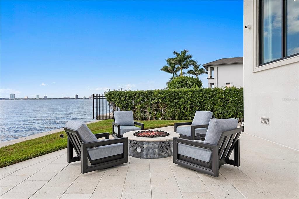 This is an inviting outdoor patio area featuring four modern armchairs arranged around a stylish fire pit. The patio is paved with light-colored tiles and overlooks a serene body of water, with a manicured lawn separating the patio from the water's edge. Lush greenery provides privacy, and the scene is set against a clear blue sky, creating a relaxing and luxurious ambiance.