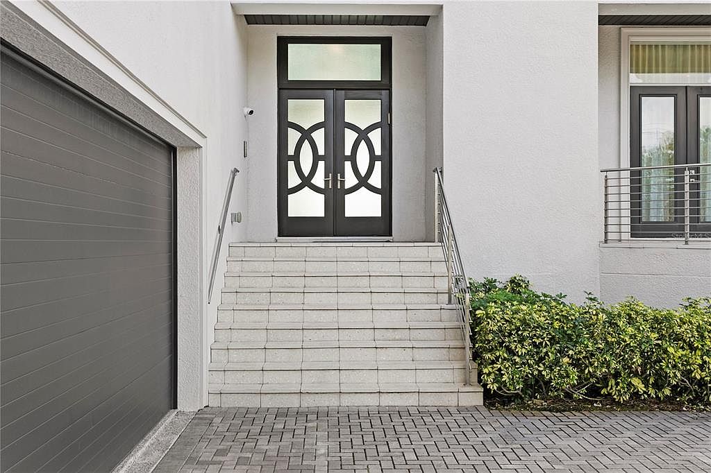 This image showcases the entryway of a modern home, featuring a set of double doors with a unique circular design and a transom window above. The entrance is accessed by a wide set of tiled stairs with stainless steel handrails, leading to a paved walkway. Lush green bushes flank the stairs, adding a touch of nature to the clean, contemporary aesthetic.