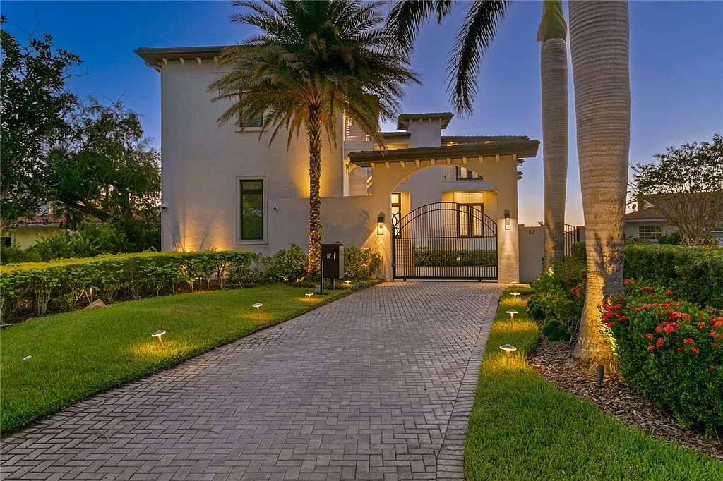 This is a front exterior view of a luxurious two-story home at dusk. The property features a brick driveway leading to an arched entryway with a wrought iron gate, lush landscaping with palm trees, and strategically placed lighting that highlights the architectural details and manicured lawn. The overall impression is one of elegance and curb appeal.
