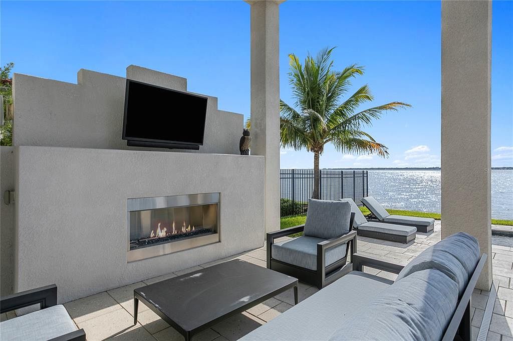 This is a luxurious outdoor patio area featuring a modern fireplace with a mounted television above it. The patio is furnished with comfortable seating, including a sofa and armchairs, and overlooks a serene waterfront view. A palm tree adds a tropical touch to the scene, enhancing the overall appeal of this outdoor living space.