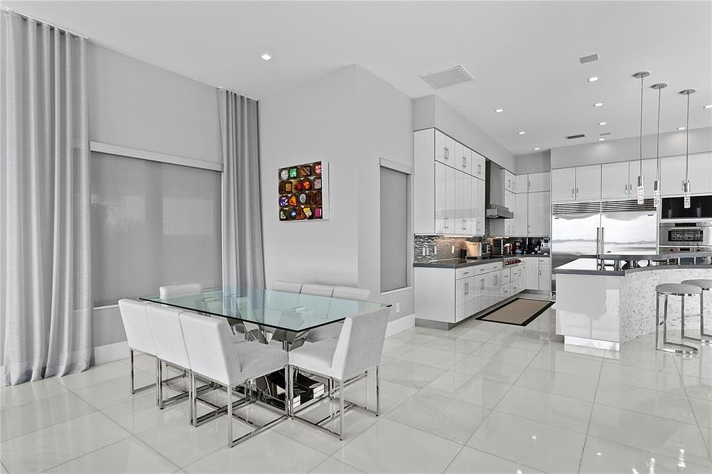 This is a bright, modern kitchen and dining area featuring white cabinetry, stainless steel appliances, and a glass-top dining table with white chairs. The kitchen island has a textured surface and bar seating. The room is well-lit with recessed lighting and pendant lights, and the white tile flooring adds to the clean, contemporary aesthetic.
