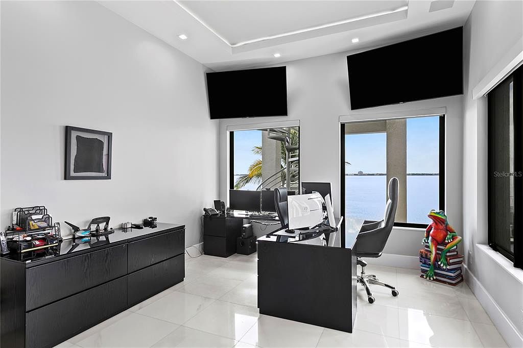 This is a modern office space featuring a sleek black desk and cabinetry, complemented by two large flat-screen TVs mounted on the wall. The room offers a view of the water through two windows, and a whimsical frog statue sits beside one of them. The overall impression is one of sophistication and functionality, ideal for a professional seeking a stylish workspace.