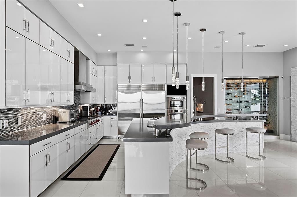 This is a modern, high-end kitchen featuring sleek white cabinetry, stainless steel appliances, and a dark countertop. A curved island with bar seating and pendant lighting adds a stylish touch. A wine cellar is visible in the background, enhancing the luxurious feel of the space.