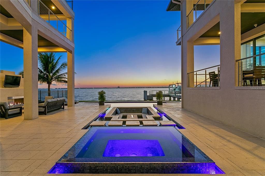 This image showcases a luxurious outdoor space featuring a modern pool and spa area with integrated fire pit. The pool is illuminated with blue lighting, creating a vibrant ambiance. The area is surrounded by a tiled patio, with views of the waterfront and a contemporary architectural design, suggesting a high-end property.