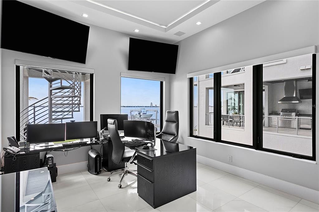 This is a modern office space featuring a sleek, dark-toned desk setup with multiple monitors and ergonomic chairs. Large windows offer views of the water and surrounding architecture, while two wall-mounted televisions add a contemporary touch. The room is well-lit with recessed lighting and natural light, creating a professional and comfortable work environment.