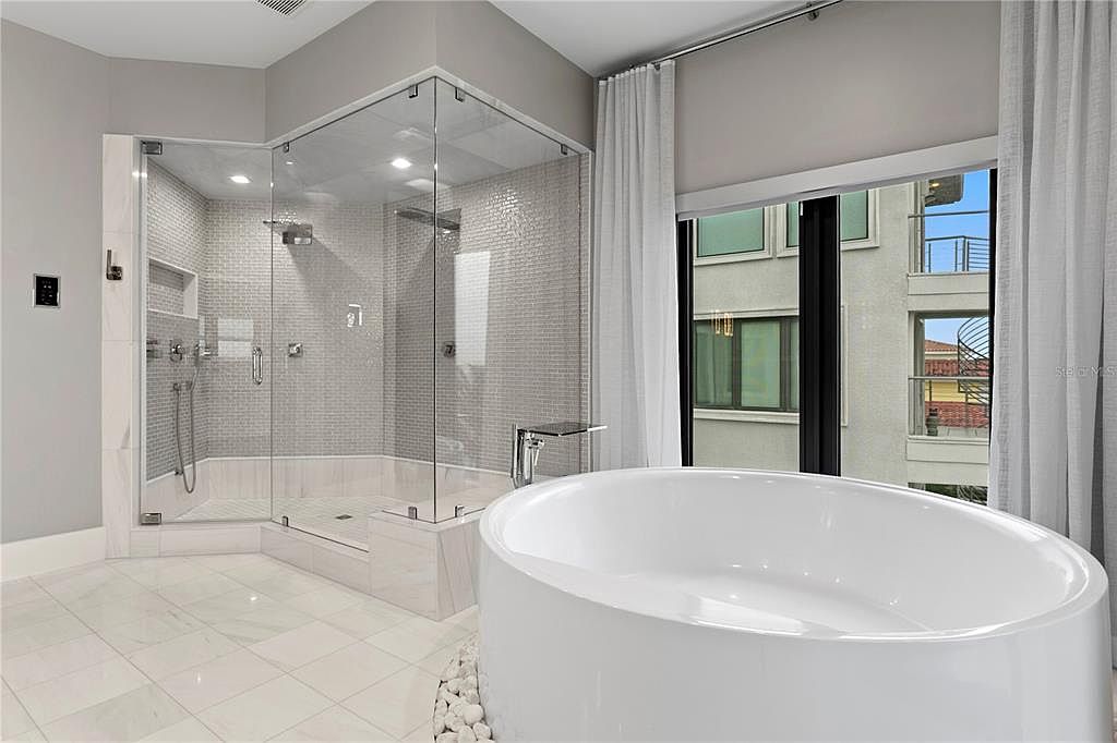 This is a luxurious primary bathroom featuring a modern, freestanding white bathtub and a glass-enclosed shower with gray mosaic tile. The bathroom has white marble flooring and is illuminated by recessed lighting. Large windows with sheer white curtains provide natural light and a view of the exterior.