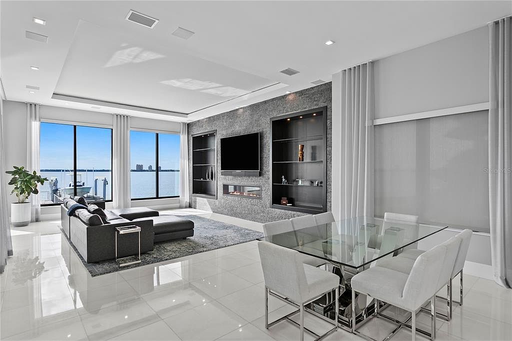 This is a bright and modern living room with a stunning water view. The room features a large sectional sofa, a glass dining table with white chairs, and a sleek entertainment center with a fireplace. The polished tile floors and neutral color palette create a luxurious and inviting atmosphere.