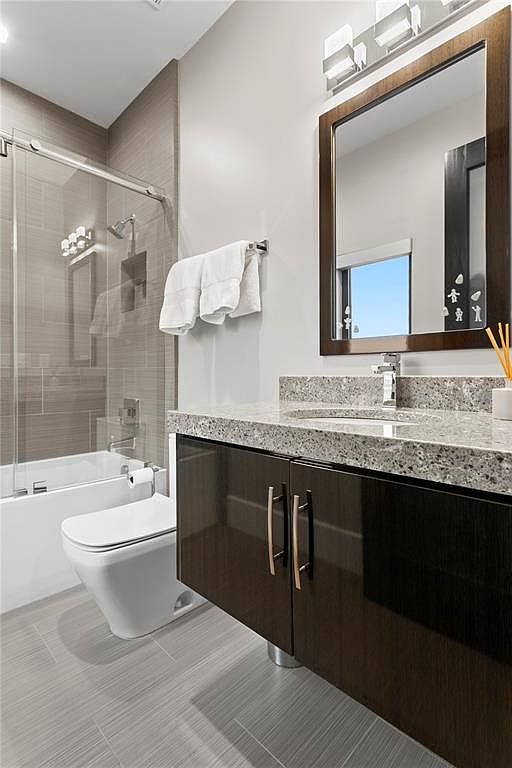 This is a well-lit bathroom featuring a modern vanity with dark wood cabinetry and a speckled granite countertop. A large mirror hangs above the sink, reflecting natural light. The bathroom includes a toilet and a bathtub with a glass shower screen, all set against a backdrop of neutral-toned tiles, creating a clean and contemporary aesthetic.