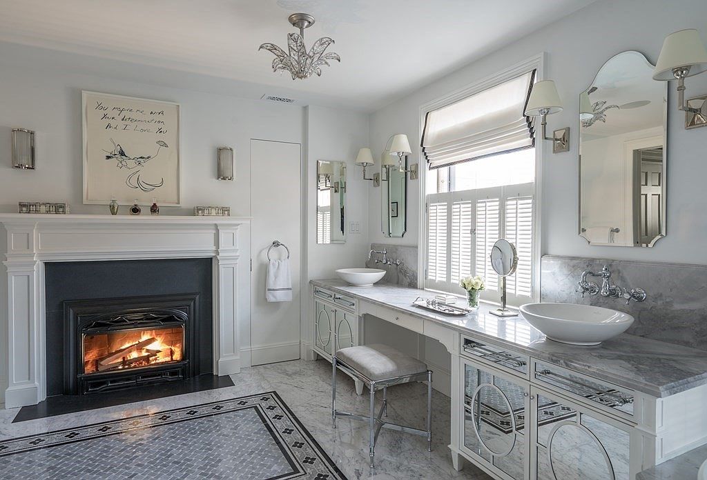 This is a luxurious primary bathroom featuring a double vanity with marble countertops and mirrored details. A fireplace adds warmth and ambiance to the space, while the chandelier provides elegant lighting. The floor is tiled with a decorative pattern, enhancing the room's sophistication.